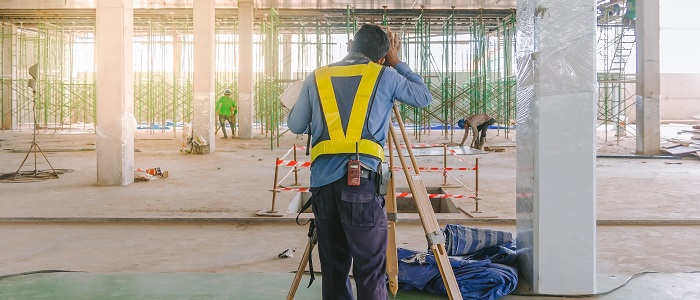 Ein Mann ist auf einer Baustelle und benutzt einen Tachymeter für eine Vermessung. Er trägt eine Sicherheitsweste.
